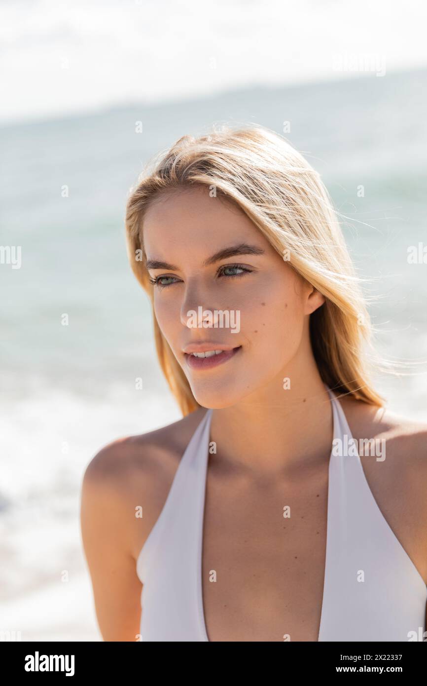 A young, beautiful blonde woman in a white bikini stands gracefully on Miami beach, embracing the tranquil scenery. Stock Photo