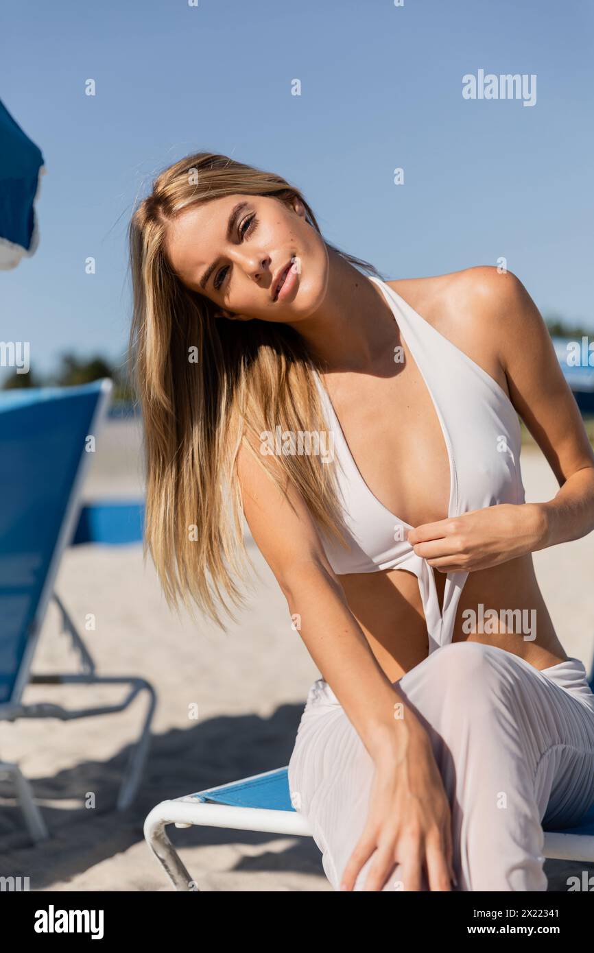 Young, beautiful blonde woman in a white bikini sitting on a beach chair, enjoying the sun and serenity of Miami beach. Stock Photo