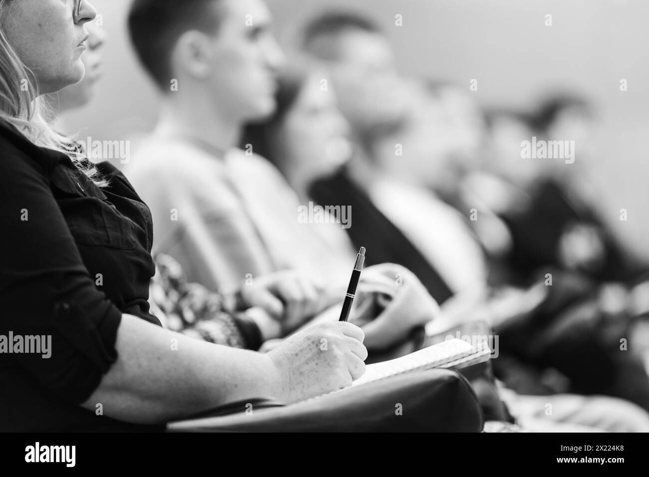 Female hands holding pen and notebook, making notes at conference lecture. Event participants in conference hall. Black and white image Stock Photo