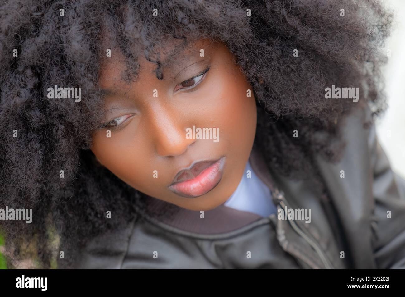 A close-up portrait captures a Black woman with voluminous natural afro hair and a subtle gaze. The soft focus on her features contrasts with her piercing eyes, expressing introspection. She's wearing a classic leather jacket, hinting at a cool, urban style. Close-Up of Contemplative Black Woman with Natural Hair. High quality photo Stock Photo