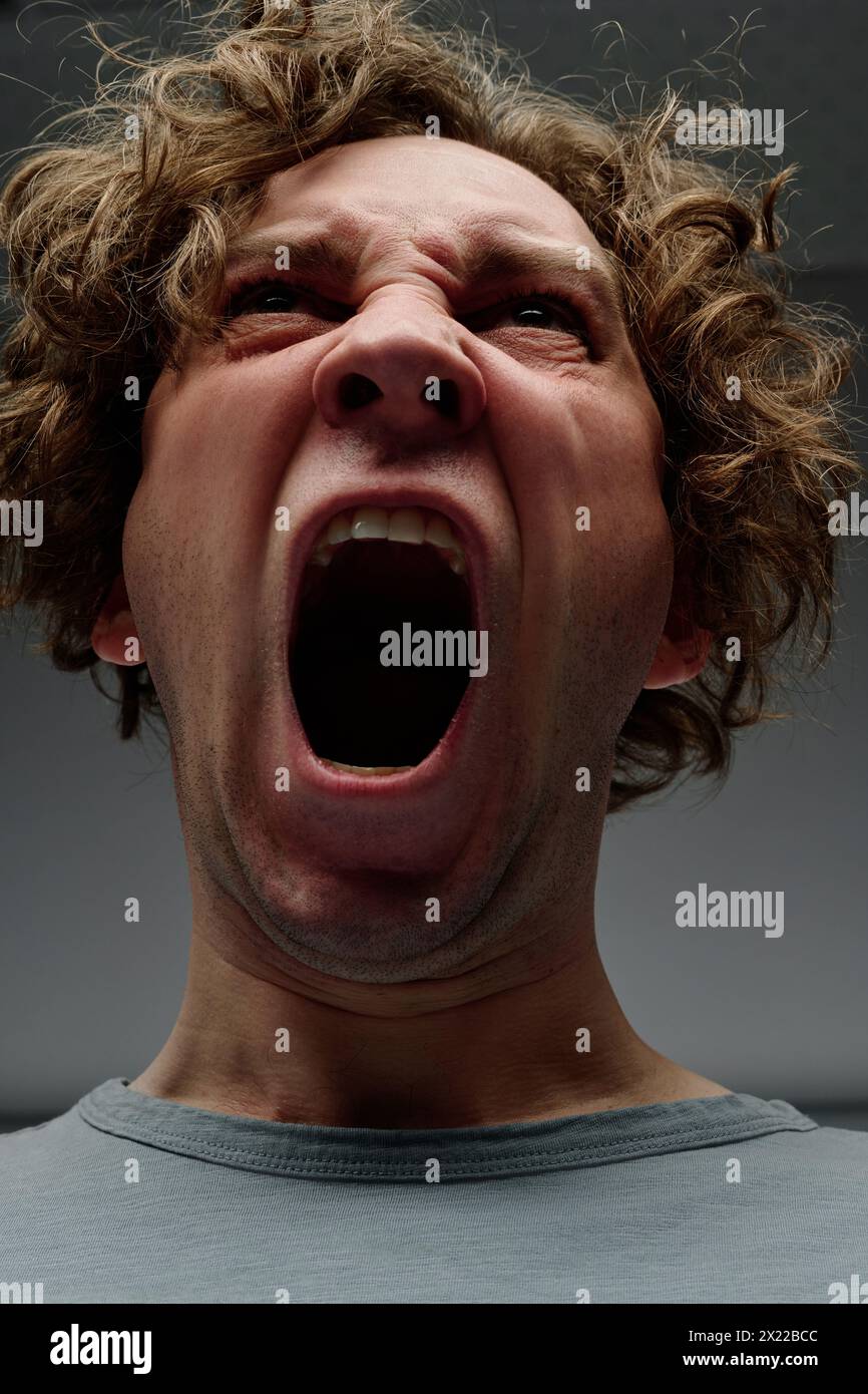 Low angle closeup studio portrait of enraged young Caucasian man with curly hair yelling Stock Photo