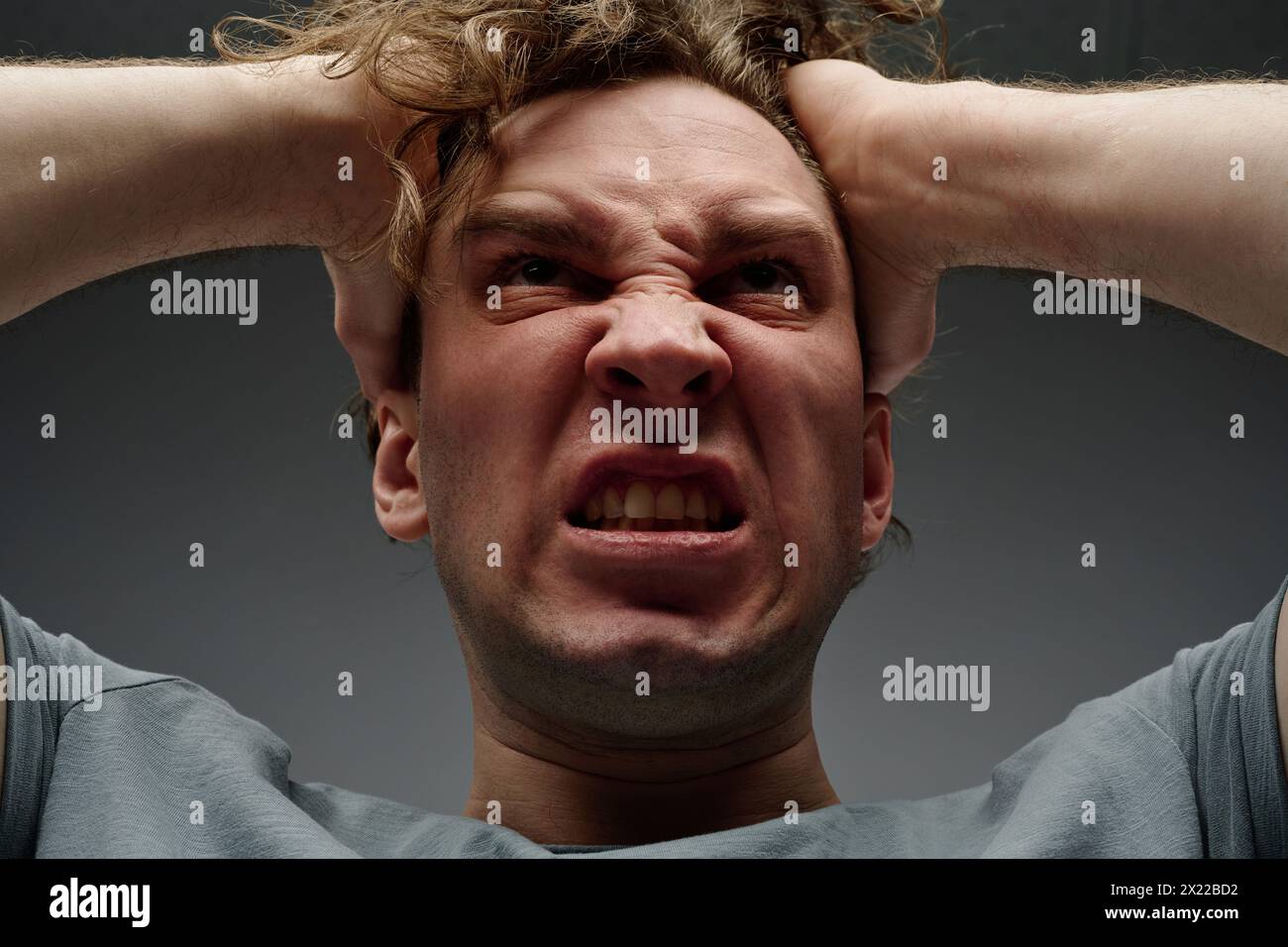Low angle closeup studio portrait of young Caucasian man expressing feeling of wrath, gray background Stock Photo
