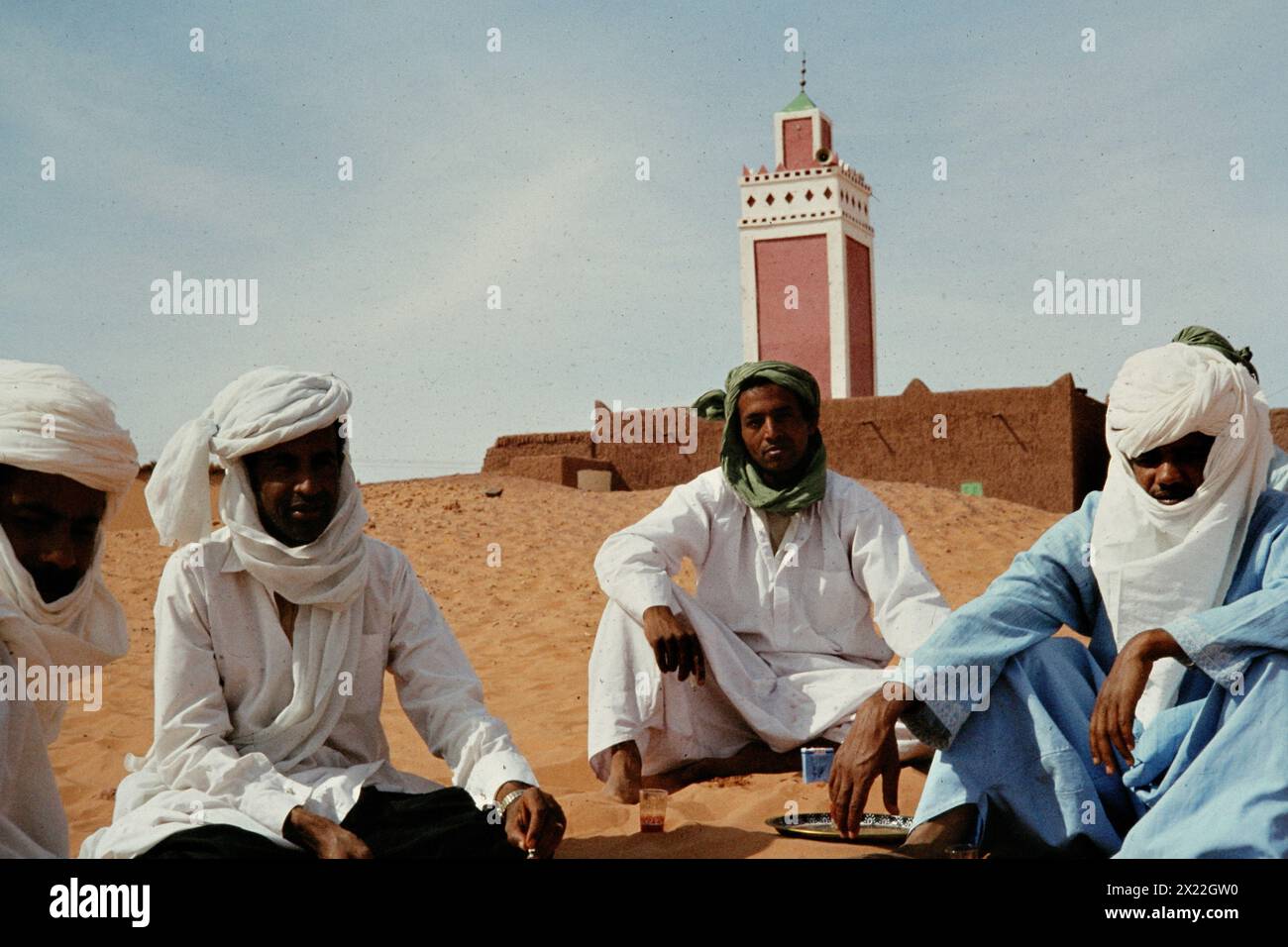 Men in traditional Tuareg clothing with facial veils sit in the sand in front of a mosque in the Algerian oasis city of Ain Salah. Stock Photo