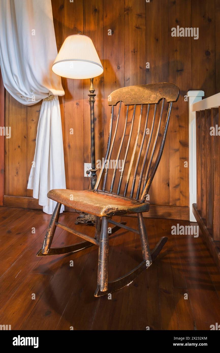 Spindle high back antique wooden rocking chair and lit pedestal lamp in hallway on upper floor inside a New Hampton style home, Quebec, Canada Stock Photo