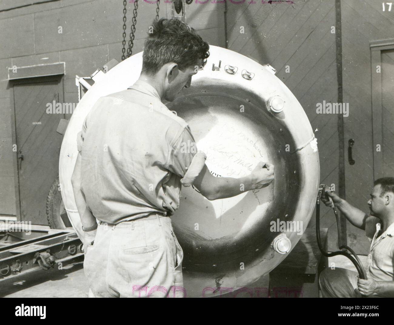 August 1945, Tinian - Scientist Dr. Norman Ramsey signing his name to Fat Man unit prior to cap being placed on the weapon. Enlisted man spraying paint on unit in background (right). Stock Photo