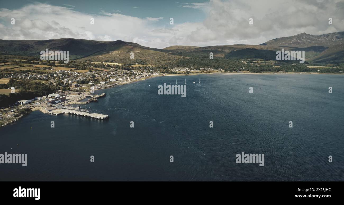 Scotland ocean port aerial view: ship, yachts and boats on coastal water of Atlantic gulf. Scottish cityscape with homes, cottages, roads against greenery hills. Cinematic shot Stock Photo