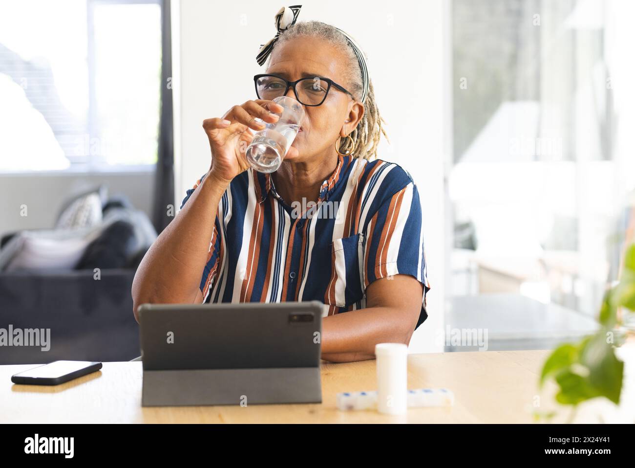 A senior African American woman drinking water at home, looking at tablet, and taking medicine Stock Photo