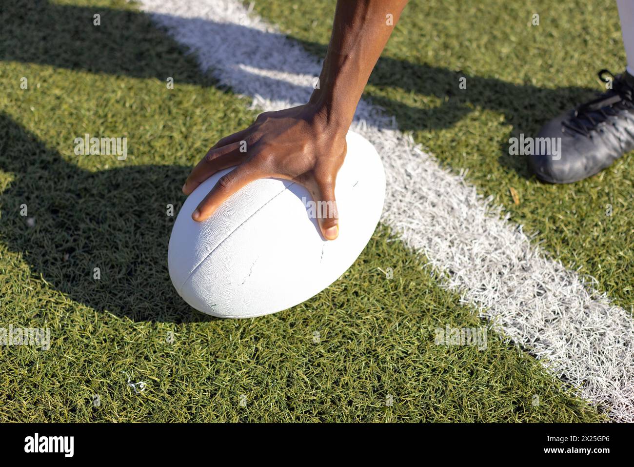 An African American young male athlete wearing cleats placing hand on rugby ball on grass field Stock Photo