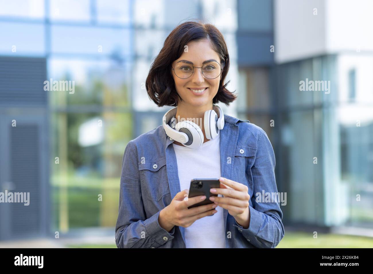 A stylish young woman in glasses and casual attire enjoys using her smartphone, with headphones around her neck, standing outdoors with a modern building background. Stock Photo