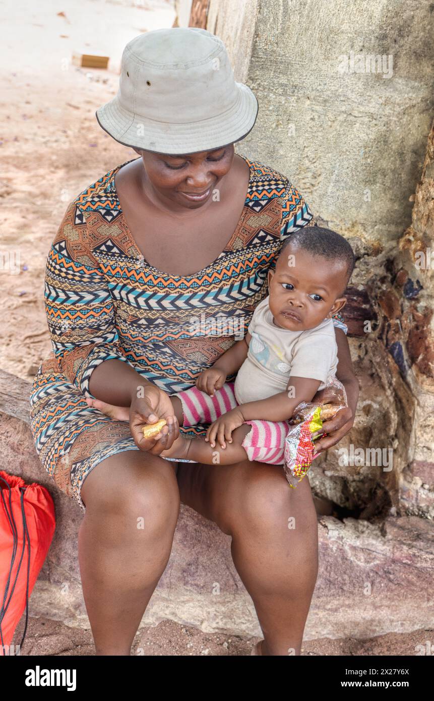 young village mother holding her hungry baby, eating some snacks, standing in front of the house Stock Photo