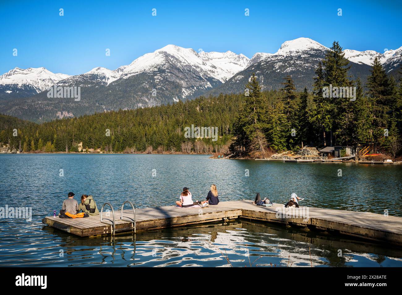 People hang out on a lake side dock in Whistler with snow covered mountains in the background.  Green Lake, Whistler BC, Canada. Stock Photo