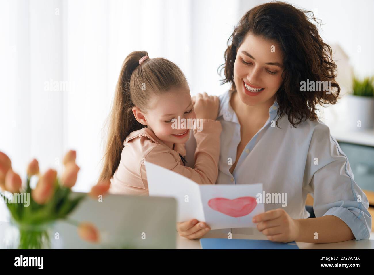 Happy mother's day. Child daughter is congratulating mom and giving her postcard. Mum and girl smiling and hugging. Family holiday and togetherness. Stock Photo