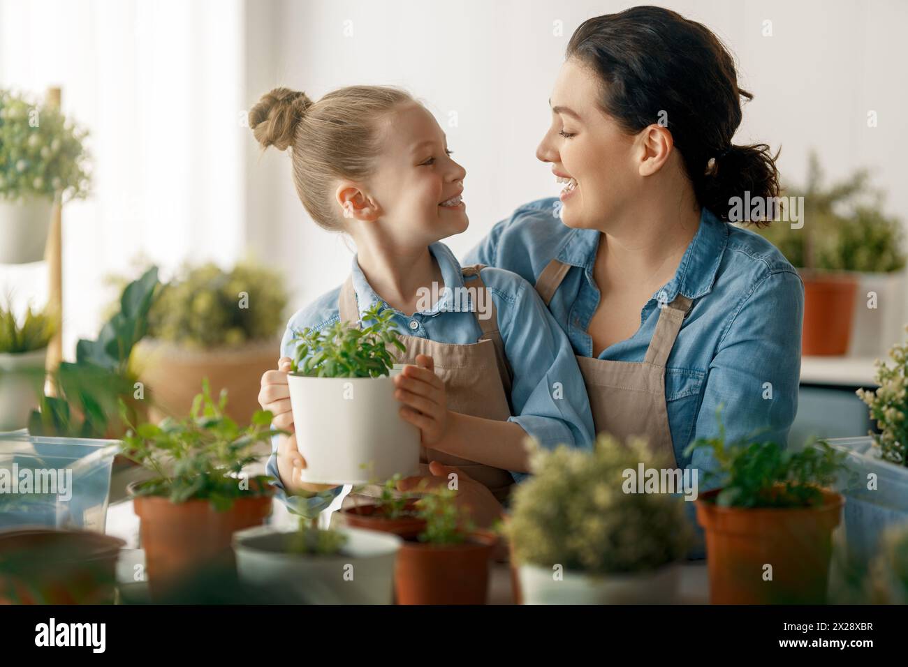 Cute child girl helping her mother to care for plants. Mom and her daughter engaging in gardening at home. Happy family in spring day. Stock Photo