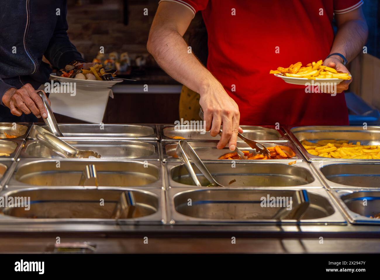 Two men help themselves to food and chips with tongs in their hands at a table with food served in stainless steel buckets at a restaurant's food buff Stock Photo