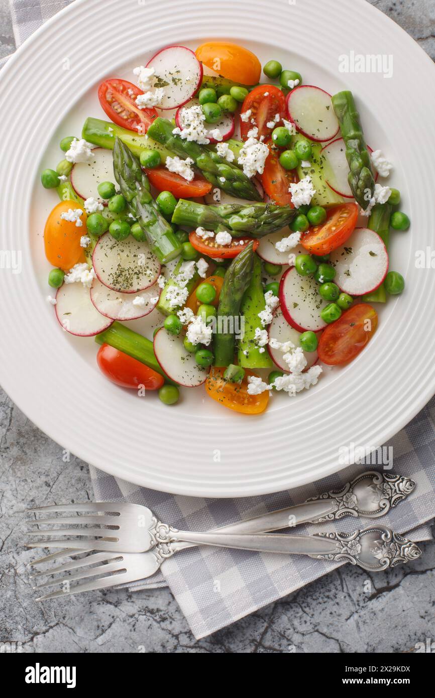 Light low-cadre salad of asparagus, fresh radishes, cherry tomatoes, green peas and goat cheese close-up in a plate on the table. Vertical top view fr Stock Photo