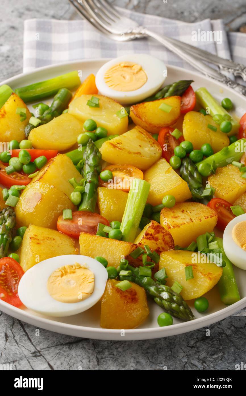 Fried potatoes with asparagus, cherry tomatoes, egg and green peas close-up in a plate on the table. Vertical Stock Photo