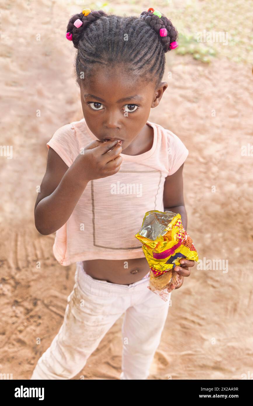 hungry village african girl with braids and beads, eating snacks in the yard Stock Photo