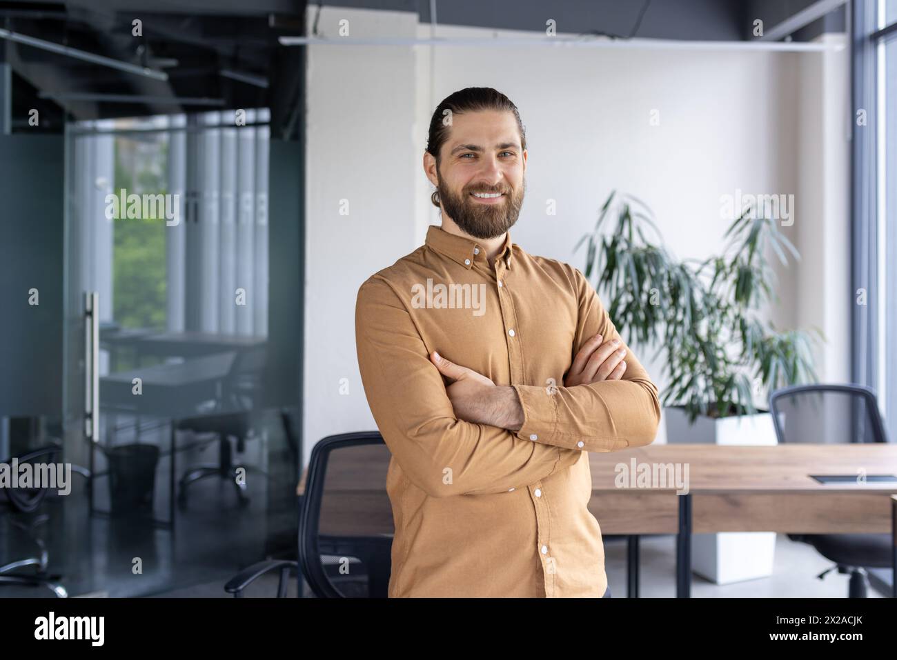 A cheerful, bearded businessman standing confidently with arms crossed in a contemporary office environment, exuding professionalism and positivity. Stock Photo