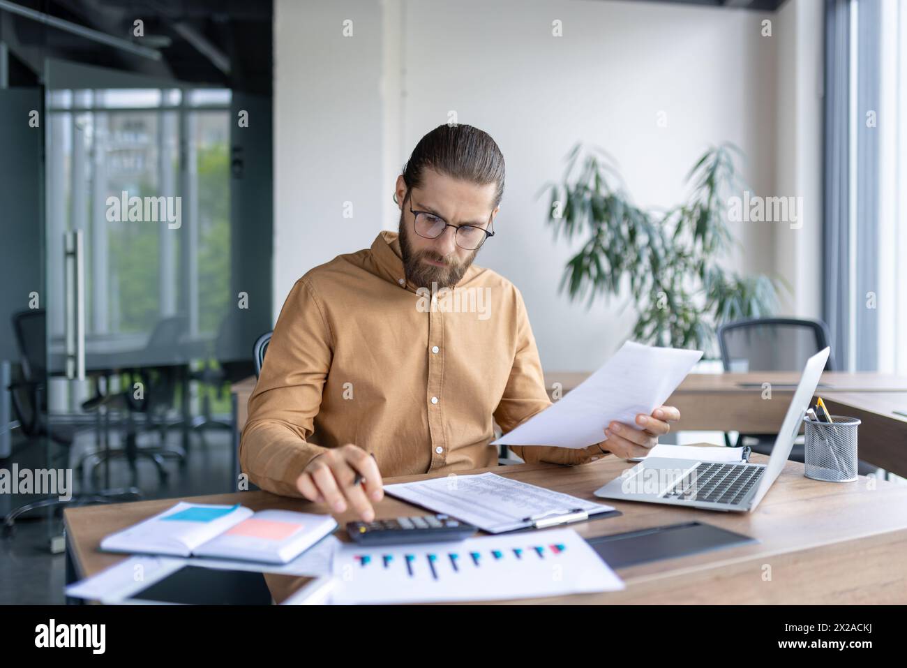 A focused young male professional examines paperwork while working in a contemporary office setting with a laptop and charts on his desk. Stock Photo