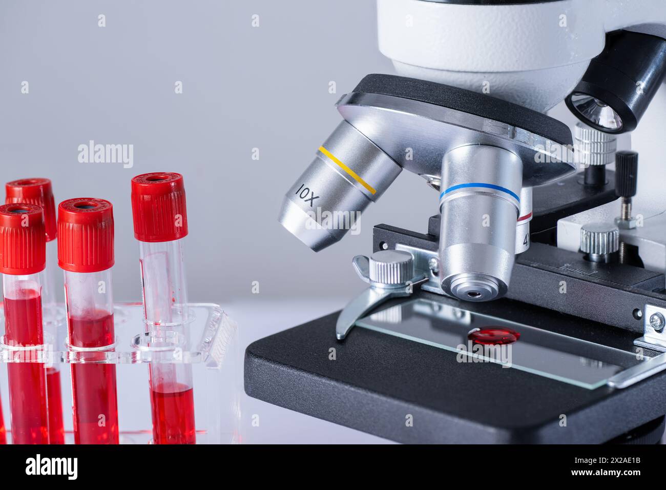 A microscope with a slide containing red blood cells. Microscope is on a laboratory table next to a few red blood cell tubes. Stock Photo