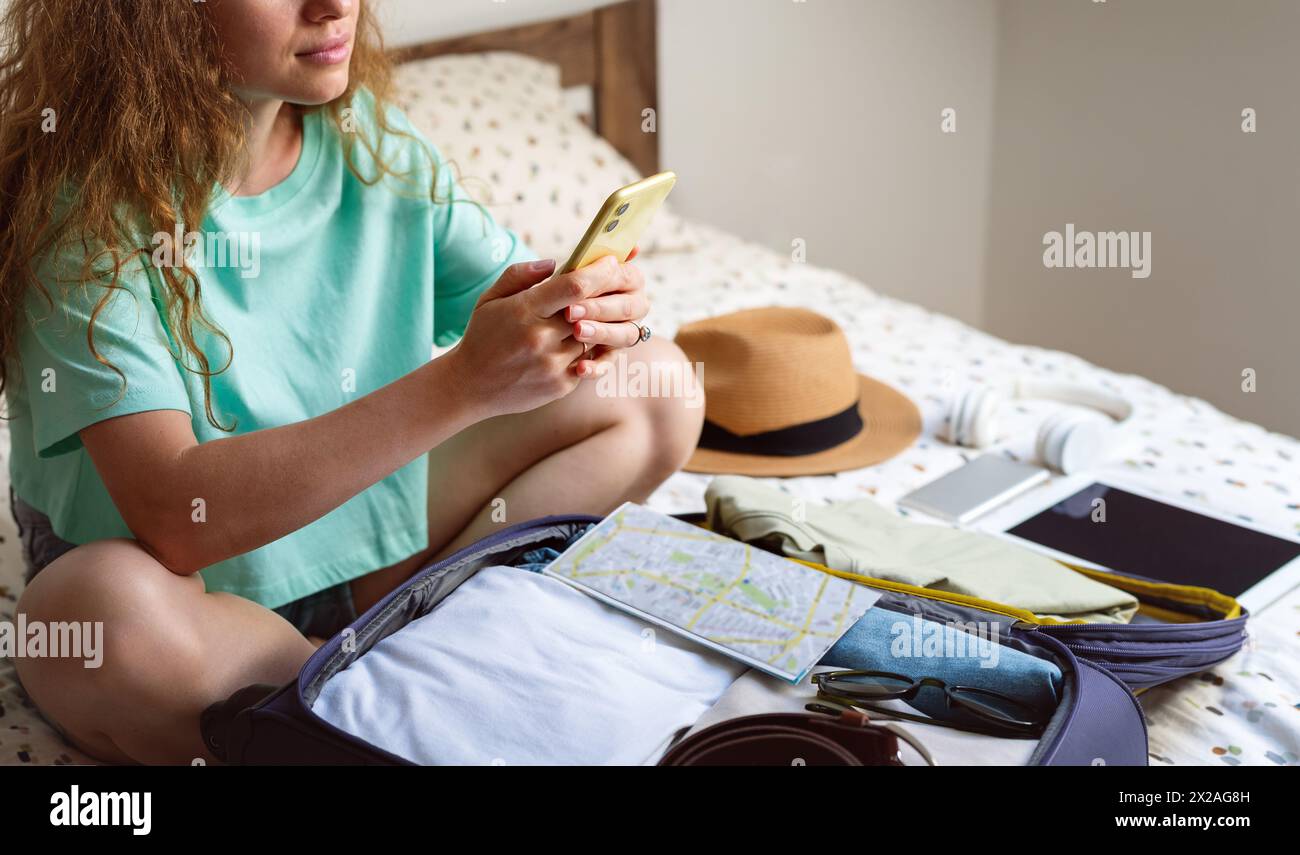 Woman packing travel bag and booking online a hotel using her smartphone. Stock Photo