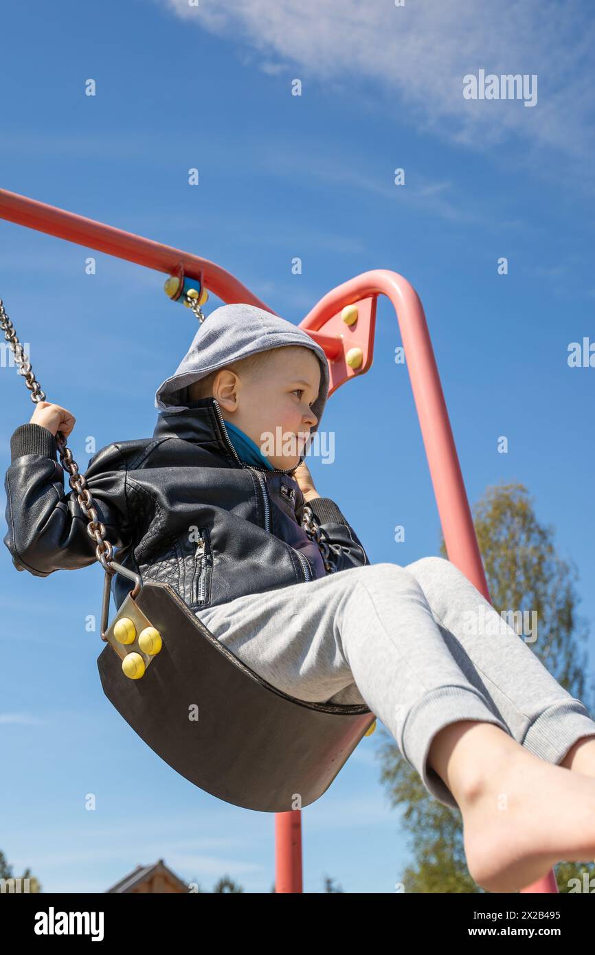 Little boy swings on a swing against a blue sky background, low angle view. Stock Photo