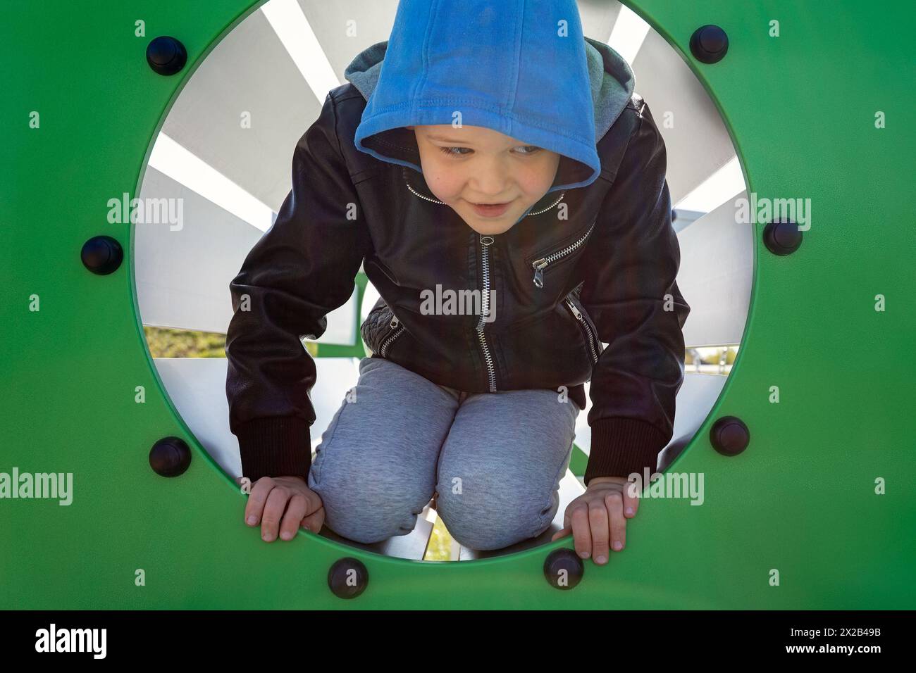 A little boy in a blue hoodie emerges from a green tube. The child performs physical exercises and trains. Stock Photo