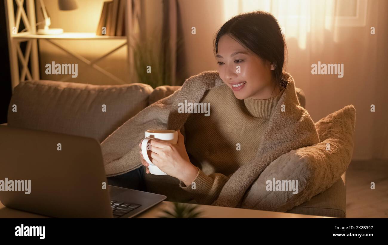 Home movie. Evening leisure. Weekend rest. Relaxed happy woman enjoying show on laptop with coffee alone on cozy couch. Stock Photo