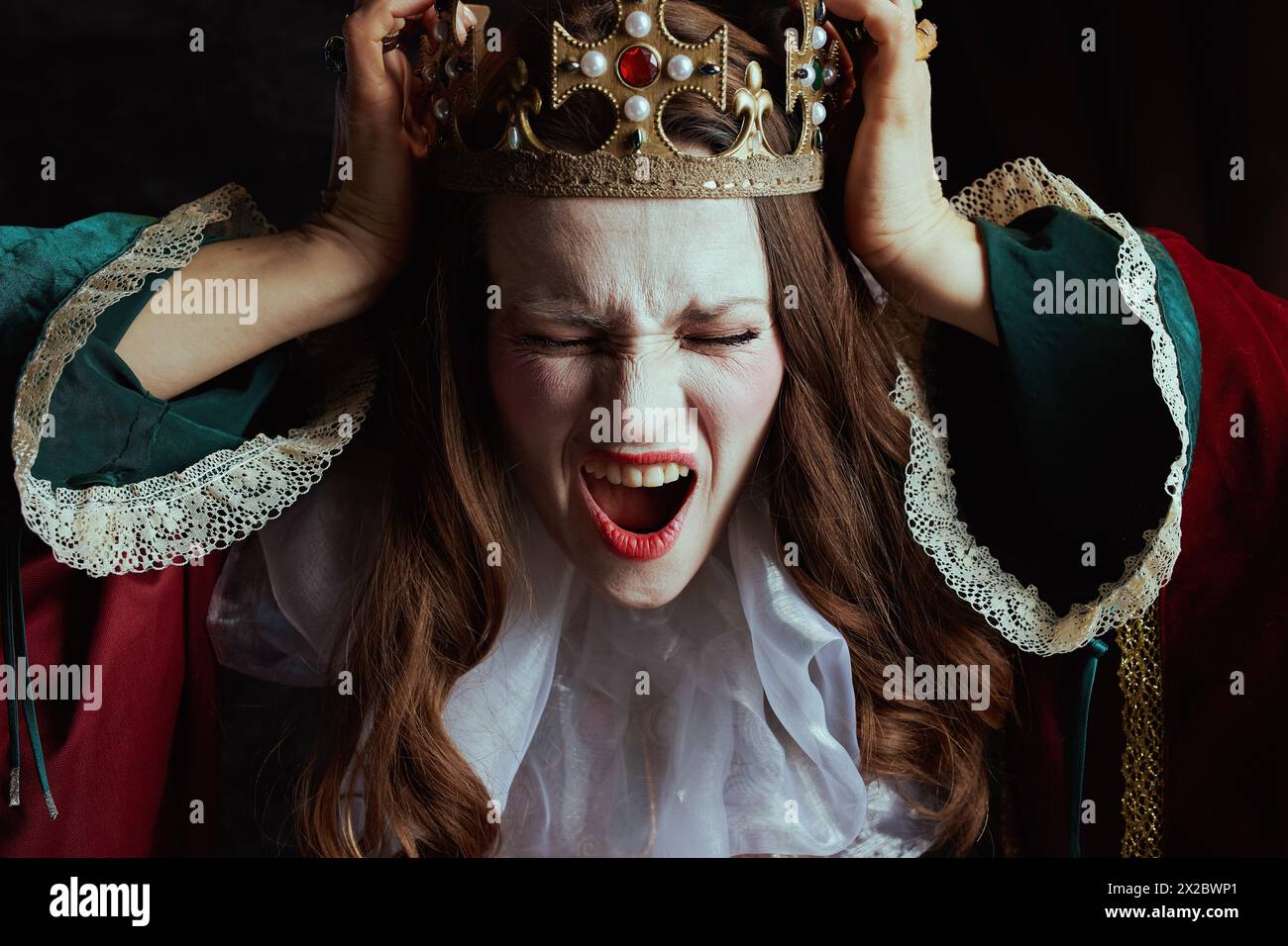 shocked medieval queen in red dress with white collar and crown on dark gray background. Stock Photo