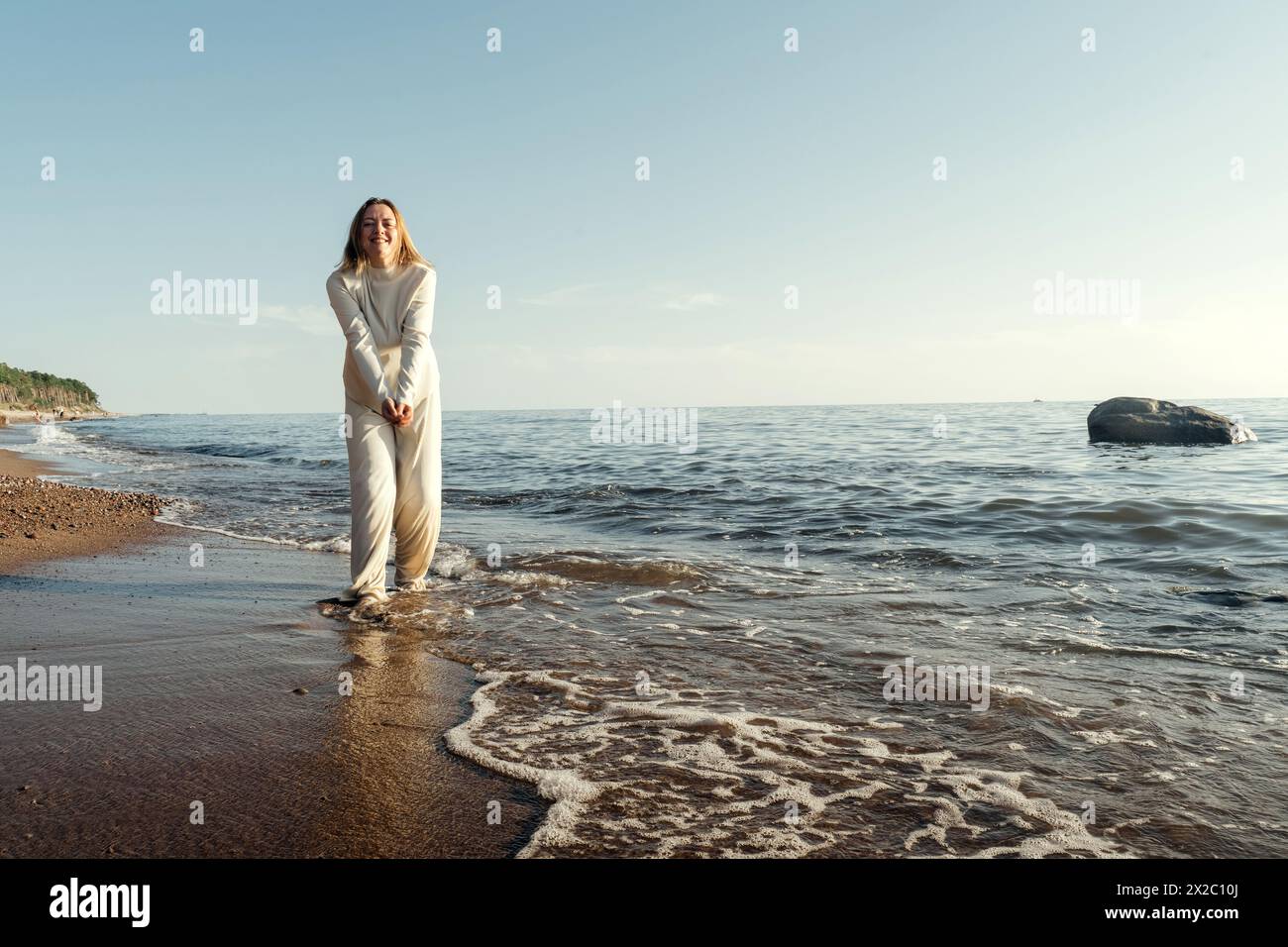 A woman is standing in the water at the beach, with waves gently lapping around her. She gazes out towards the horizon under the clear blue sky. Stock Photo