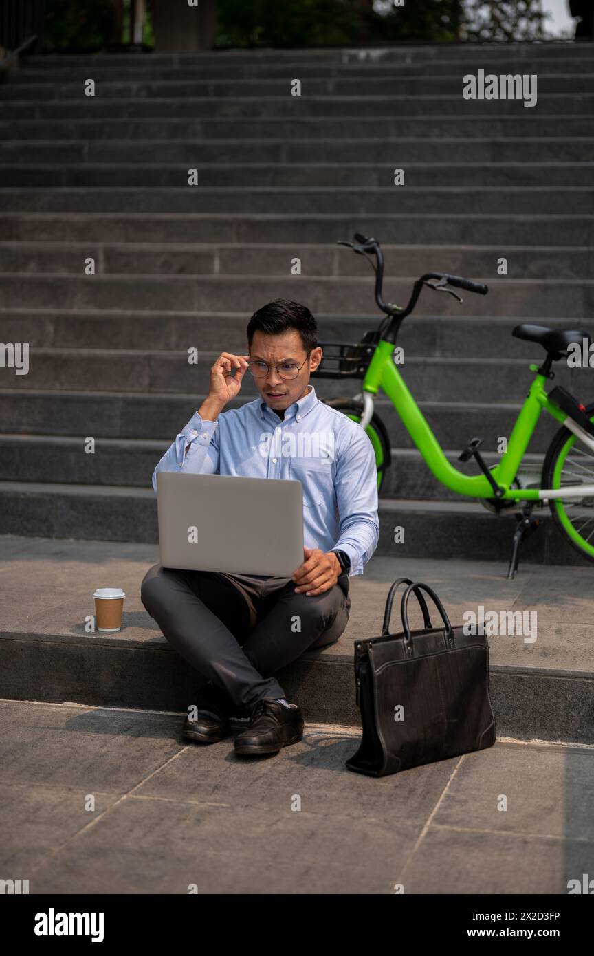 A focused Asian millennial businessman sits on outdoor steps, using his laptop, with a green bicycle parked beside him. modern work culture, city life Stock Photo