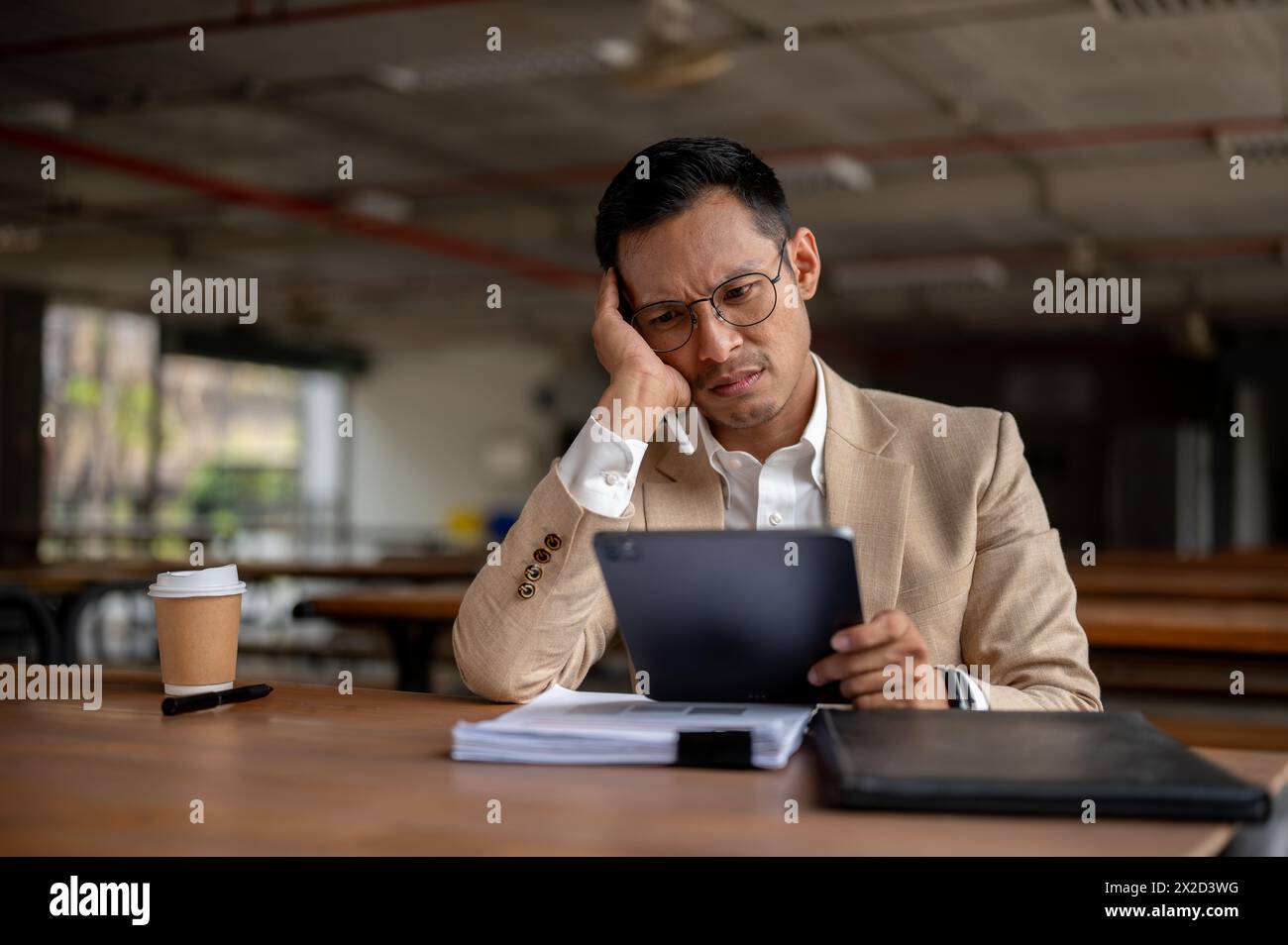 A contemplative Asian millennial businessman looks pensive while holding a digital tablet, resting his head on his hand, facing a tedious problem at w Stock Photo