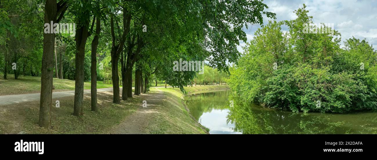 footpath in summer green park along water canal on bright sunny day. panorama. Stock Photo