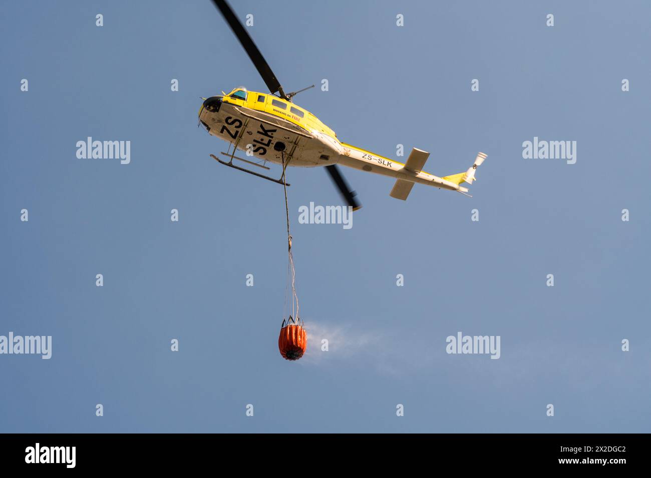 working on fire yellow emergency helicopter carrying bambi water bucket to a fire scene in Cape Town South Africa concept aerial firefighting Stock Photo