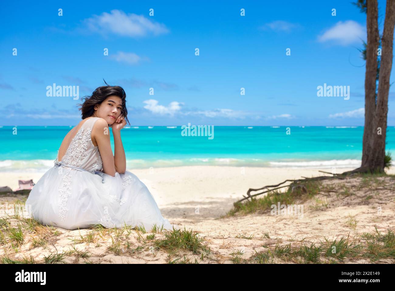 Biracial teen girl in white dress sitting by beautiful blue Hawaiian ocean white white sandy beach in background Stock Photo