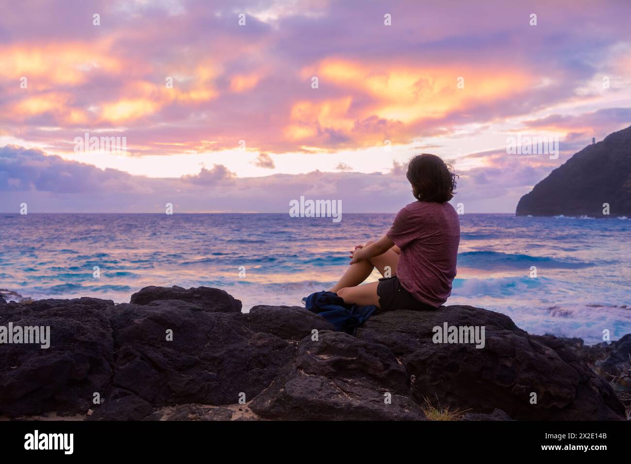 Young woman sitting on rocky beach watching the sun rise over the Hawaiian ocean at Makapu'u Beach, Oahu Stock Photo
