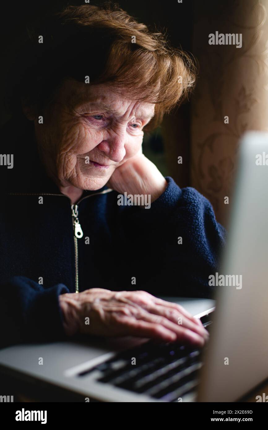 An elderly woman sits with her laptop, bathed in its soft glow, her face serene, absorbed in digital pursuits. Stock Photo