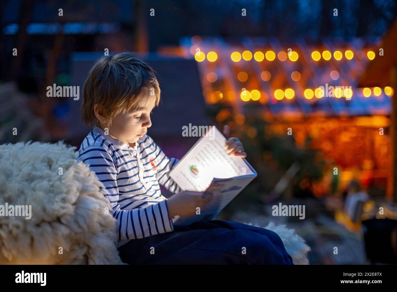 Beautiful blond child, boy, reading a book at night with flashligh, sitting on the stairs in garden in cottage village house Stock Photo