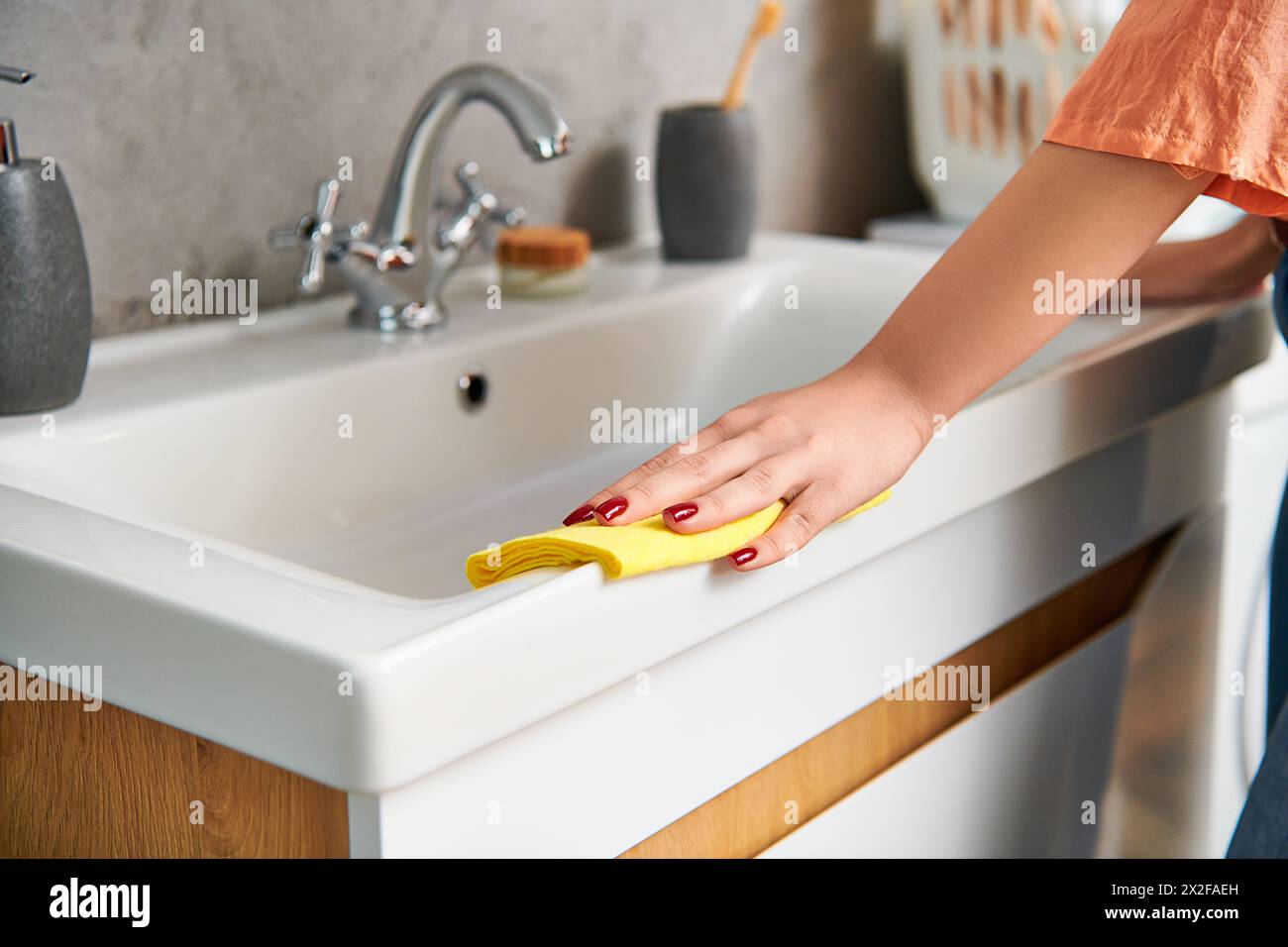 A stylish woman in casual attire diligently scrubs a sink with a bright yellow sponge to remove dirt and achieve a spotless shine. Stock Photo