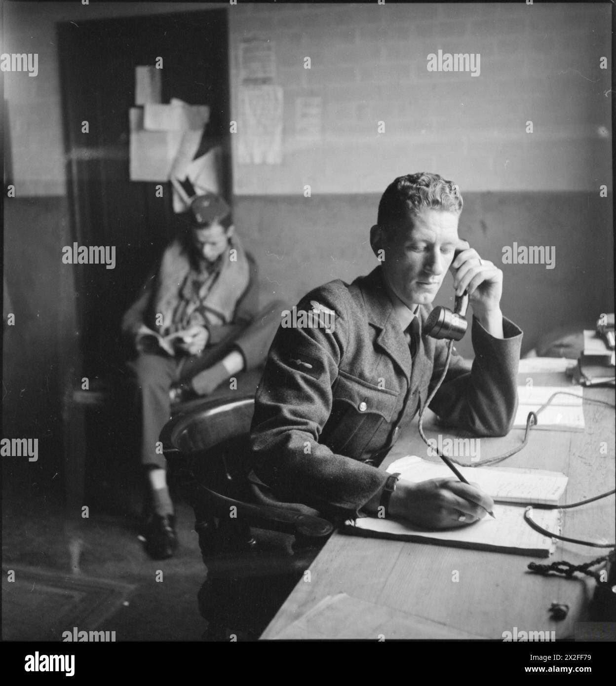 AMERICANS IN BRITAIN: THE WORK OF NO 121 (EAGLE) SQUADRON RAF, ROCHFORD, ESSEX, AUGUST 1942 - In the Dispersal Hut at Rochford airfield, the telephonist logs a call from the Control Tower, confirming that 12 Spitfires are successfully airborne. In the background, a 'spare' pilot reads a book Royal Air Force Stock Photo