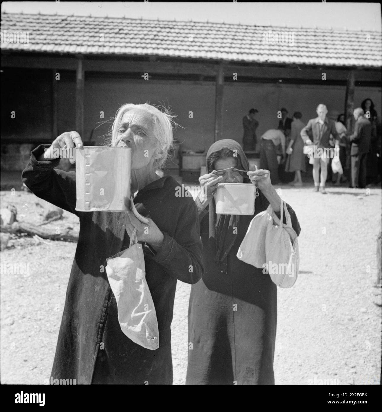 THE LIBERATION OF RHODES, 1945 - Two elderly Greek women sip their soup as they leave a Red Cross kitchen. They also carry bags so that they can call at a food distribution centre to collect their rations. During the German occupation the local inhabitants had suffered the effects of malnutrition due to insufficient food. After the liberation of the island the British quickly began to organize food supplies, issuing people with ration books and distributing essential food stuffs. Red Cross kitchens also assisted by providing nourishing soup Stock Photo