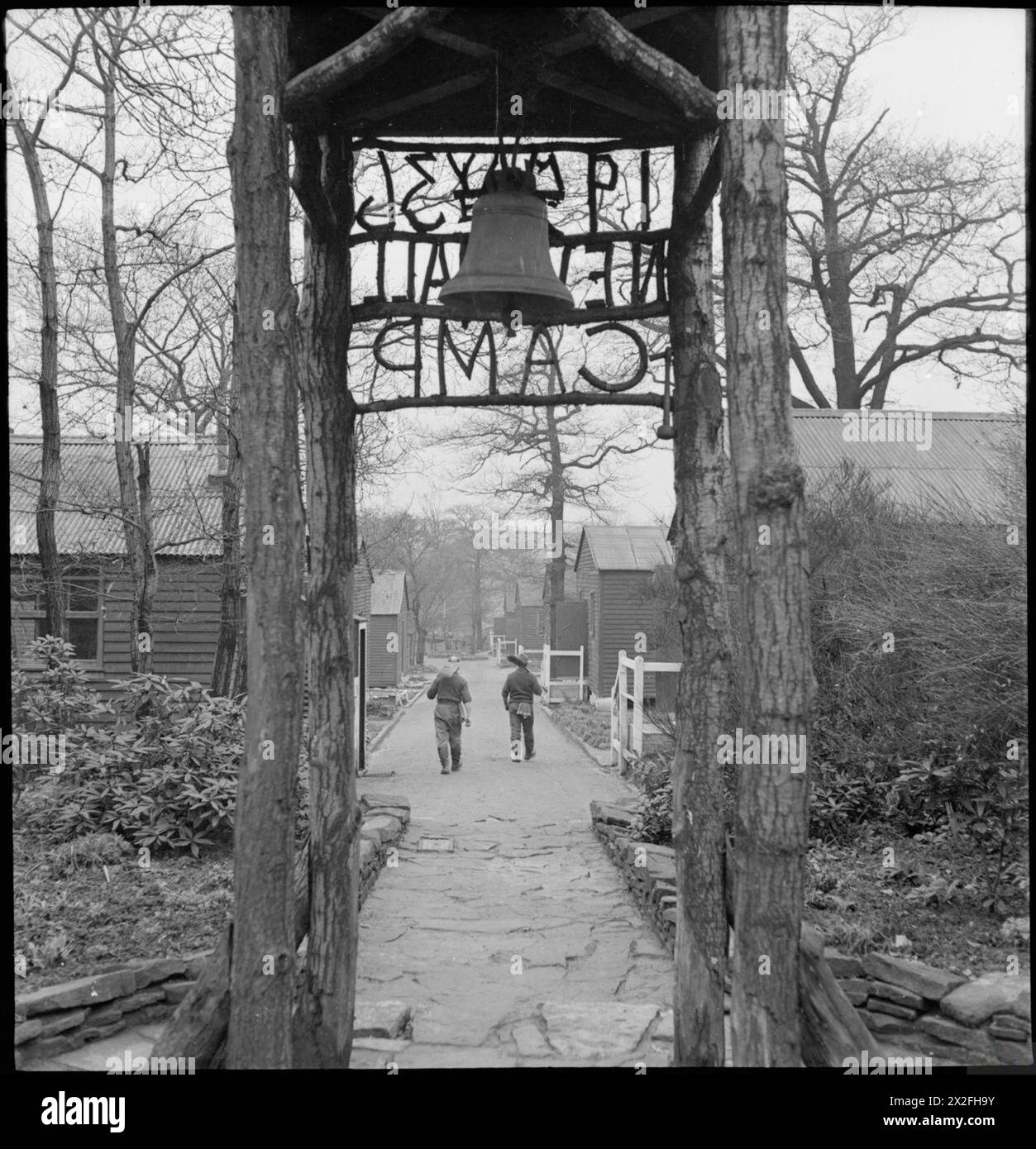 WAKEFIELD TRAINING PRISON AND CAMP: EVERYDAY LIFE IN A BRITISH PRISON, WAKEFIELD, YORKSHIRE, ENGLAND, 1944 - The entrance to New Hall Camp at Wakefield Training Prison. Two inmates walk down the path towards their huts, carrying garden tools. According to the original caption, the land for this camp was acquired in 1933. 'Parties of prisoners conveyed daily from Wakefield Prison cleared the woodland for cultivation and gradually erected the camp' Stock Photo
