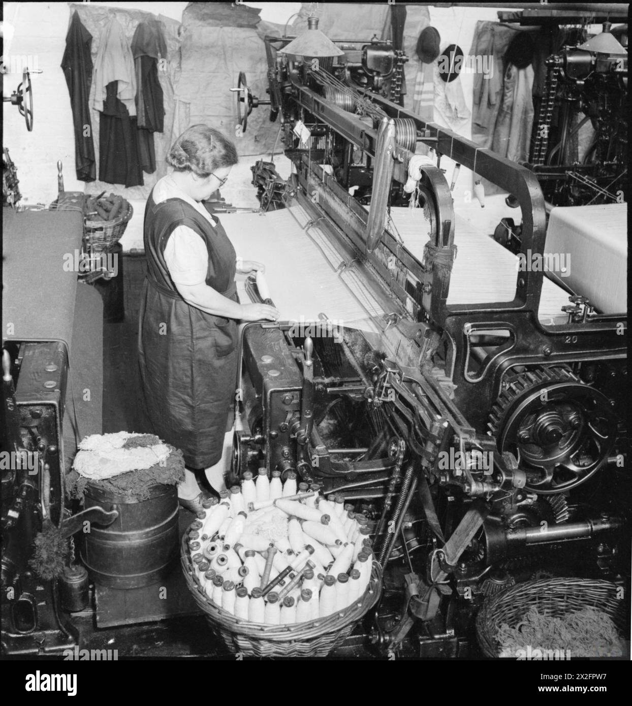 OLD RAGS INTO NEW CLOTH: SALVAGE IN BRITAIN, APRIL 1942 - A mill girl works at a loom in at textile mill, somewhere in Britain. The weaving loom intertwines separate threads of warp and weft to produce woollen cloth. The warp is the thread which runs lengthways in the machine, with the weft being shuttled back and forth through it. The mill worker pictured here is putting the weft bobbin into the shuttle of the loom. A large basket of bobbins can be seen beside her Stock Photo