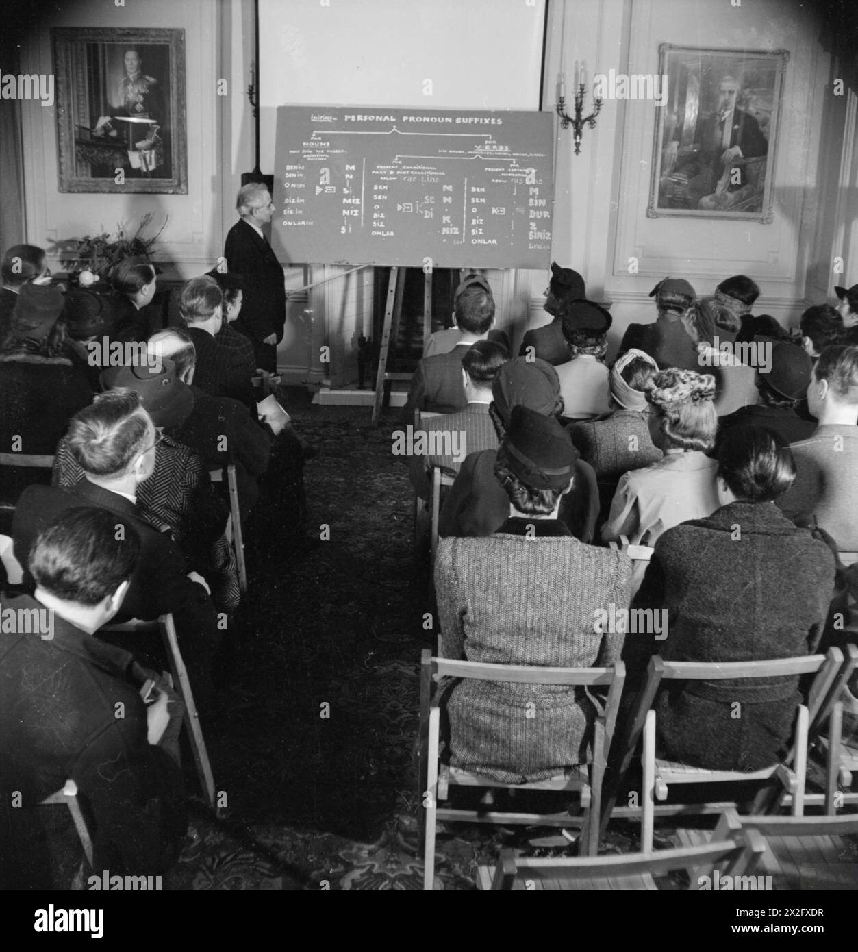 BRITONS LEARN TURKISH: ADULT EDUCATION IN LONDON, 1943 - Bay Ali Riza Sencan, Secretary of the Halkevi, points to the blackboard whilst teaching the class about the three different personal pronoun suffixes. The students sit in rows in an ornate room, with portraits of King George VI and President Inonu on display on the walls. Turkish language classes are held here twice a week Stock Photo