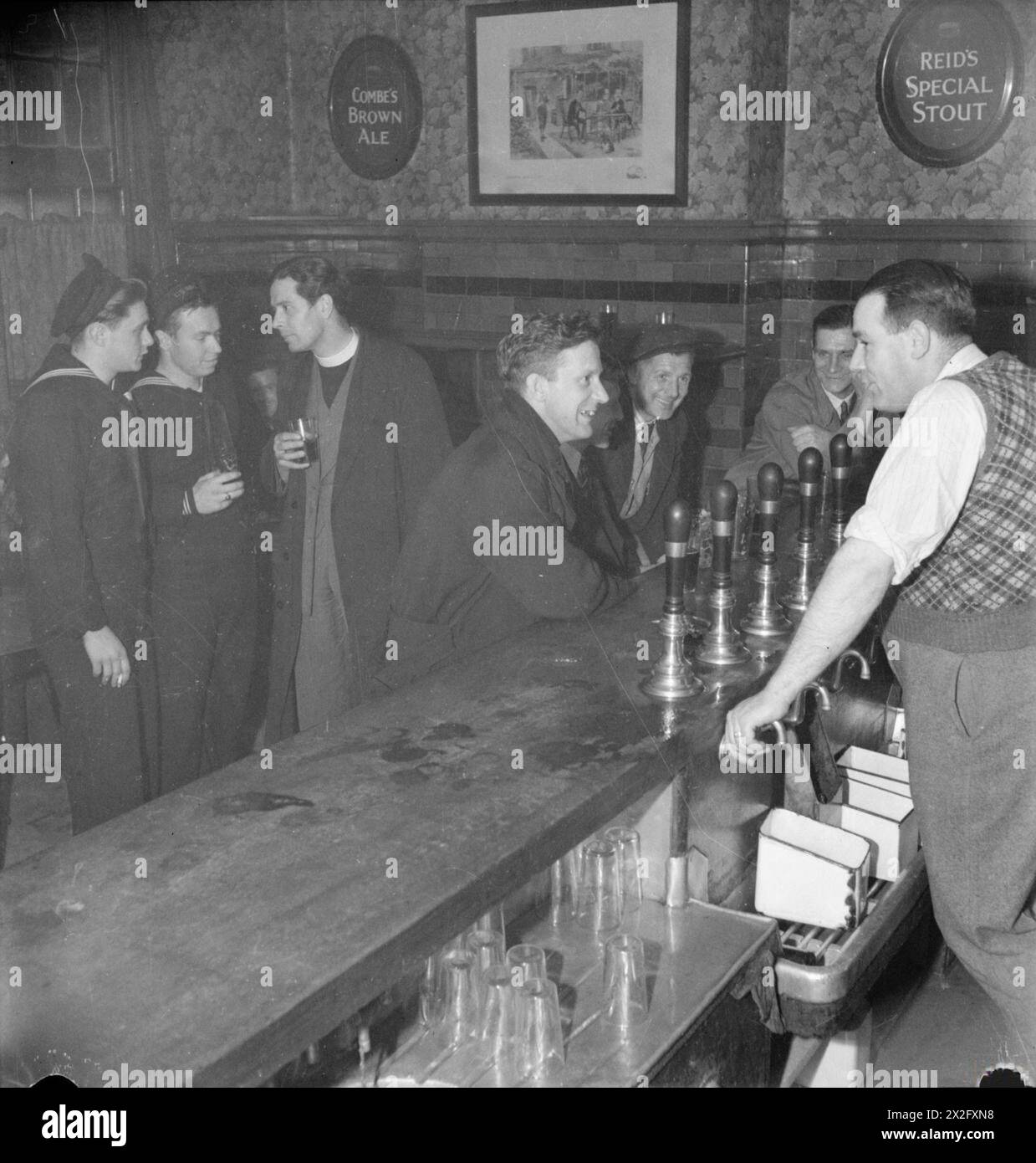 PARISH PRIEST: THE WORK OF THE VICAR OF ST MARK'S CHURCH, VICTORIA DOCKS, SILVERTOWN, LONDON, ENGLAND, UK, 1944 - Reverend Joseph Stephens chats to American sailors William Eiseneacher (of 204 East Main Street, Tremont, Pennsylvania) and John Bacher (of 1617 North Spring Street, Jeffersonville, Indiana) in the 'Royal Albert' pub in Silvertown. Several other men can be seen sitting at the bar, sharing a joke with the landlord Stock Photo