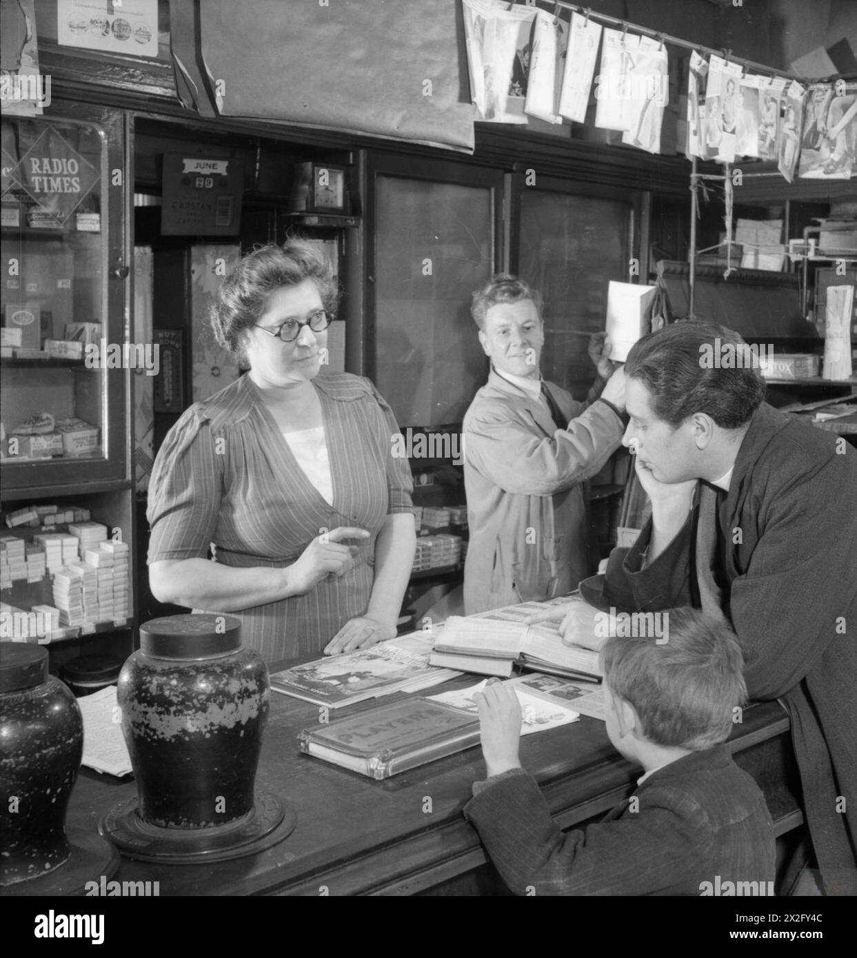 PARISH PRIEST: THE WORK OF THE VICAR OF ST MARK'S CHURCH, VICTORIA DOCKS, SILVERTOWN, LONDON, ENGLAND, UK, 1944 - Reverend Joseph Stephens runs through the music for Sunday service with newsagent and church organist Mrs Fry in her shop in Silvertown. A small boy stands next to Rev Stephens as he waits to buy stamps from Mr Fry, who is working behind the counter with his wife Stock Photo