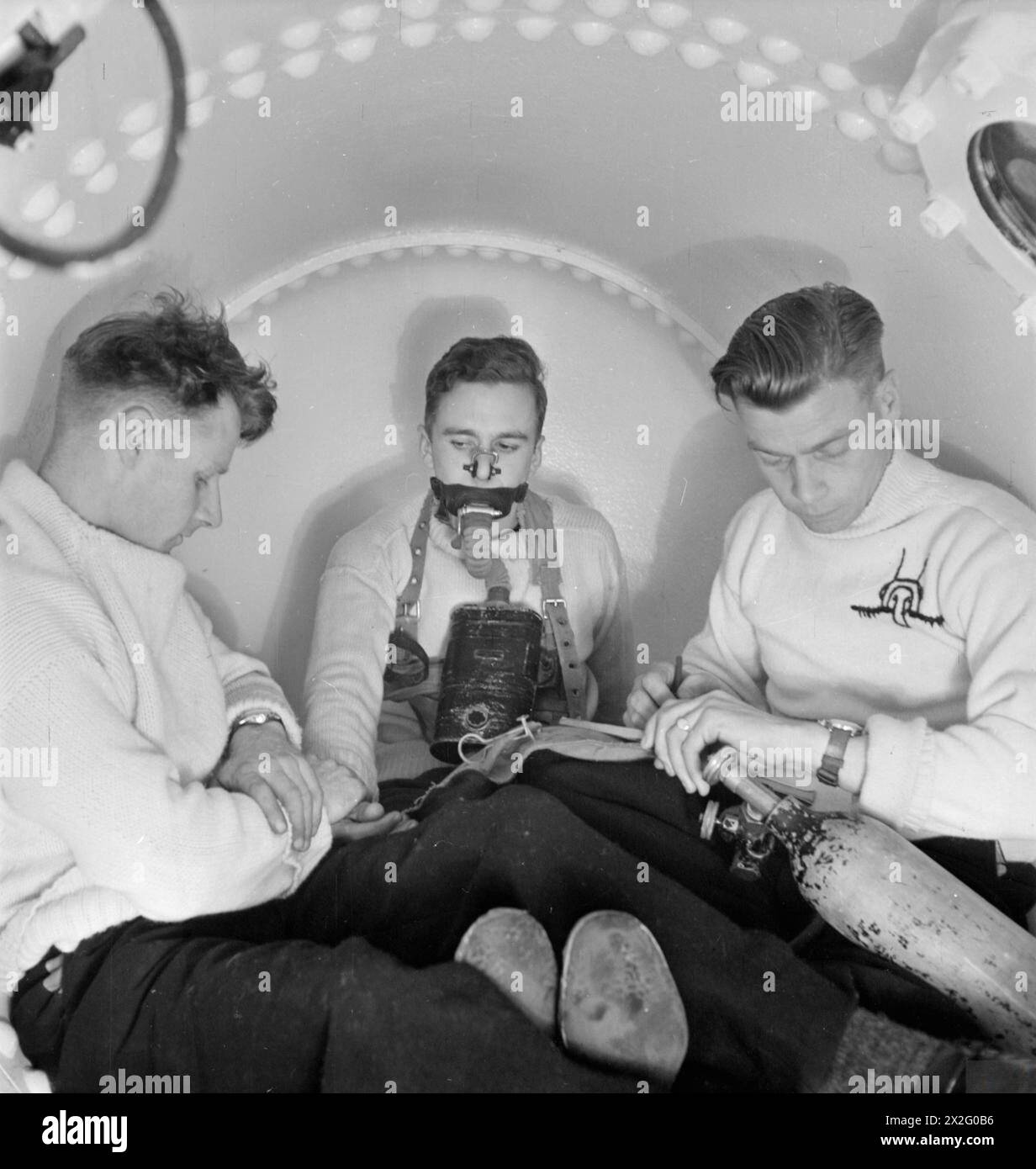 BRITISH UNDERWATER SECRETS. JUNE 1945, ADMIRALTY EXPERIMENTAL DIVING UNIT, TOLWORTH, SURREY. - An experimental subject breathing oxygen at high pressure in the compression chamber, while the chief attendant, a submarine coxswain (left) is taking his pulse. Attendant on the right is supplying gas into the apparatus and keeping guard Stock Photo