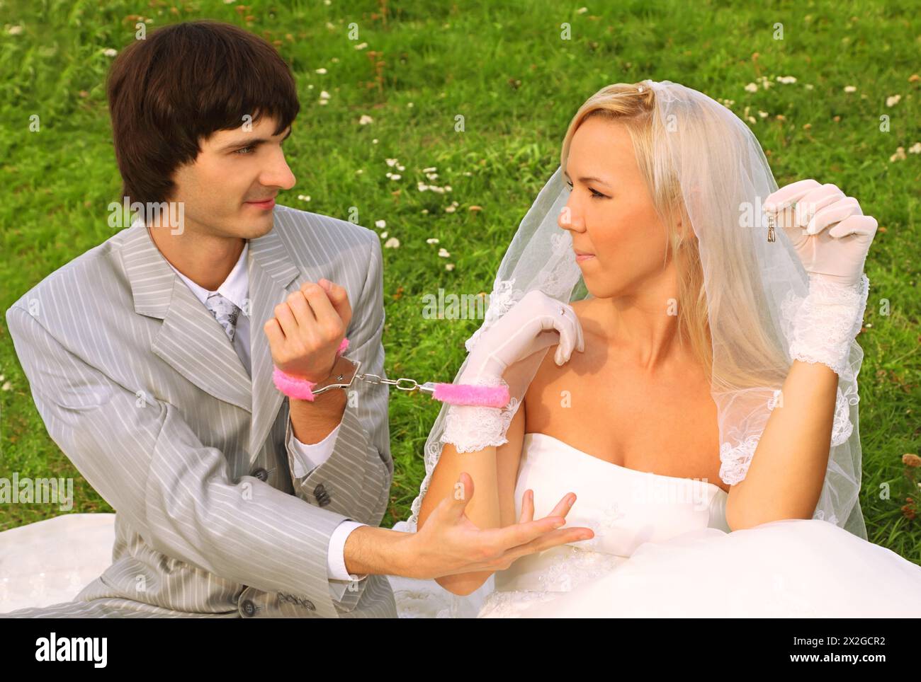 Beautiful young groom and bride wearing white dress sitting on green grass and handcuffed,  groom ask of bride key Stock Photo