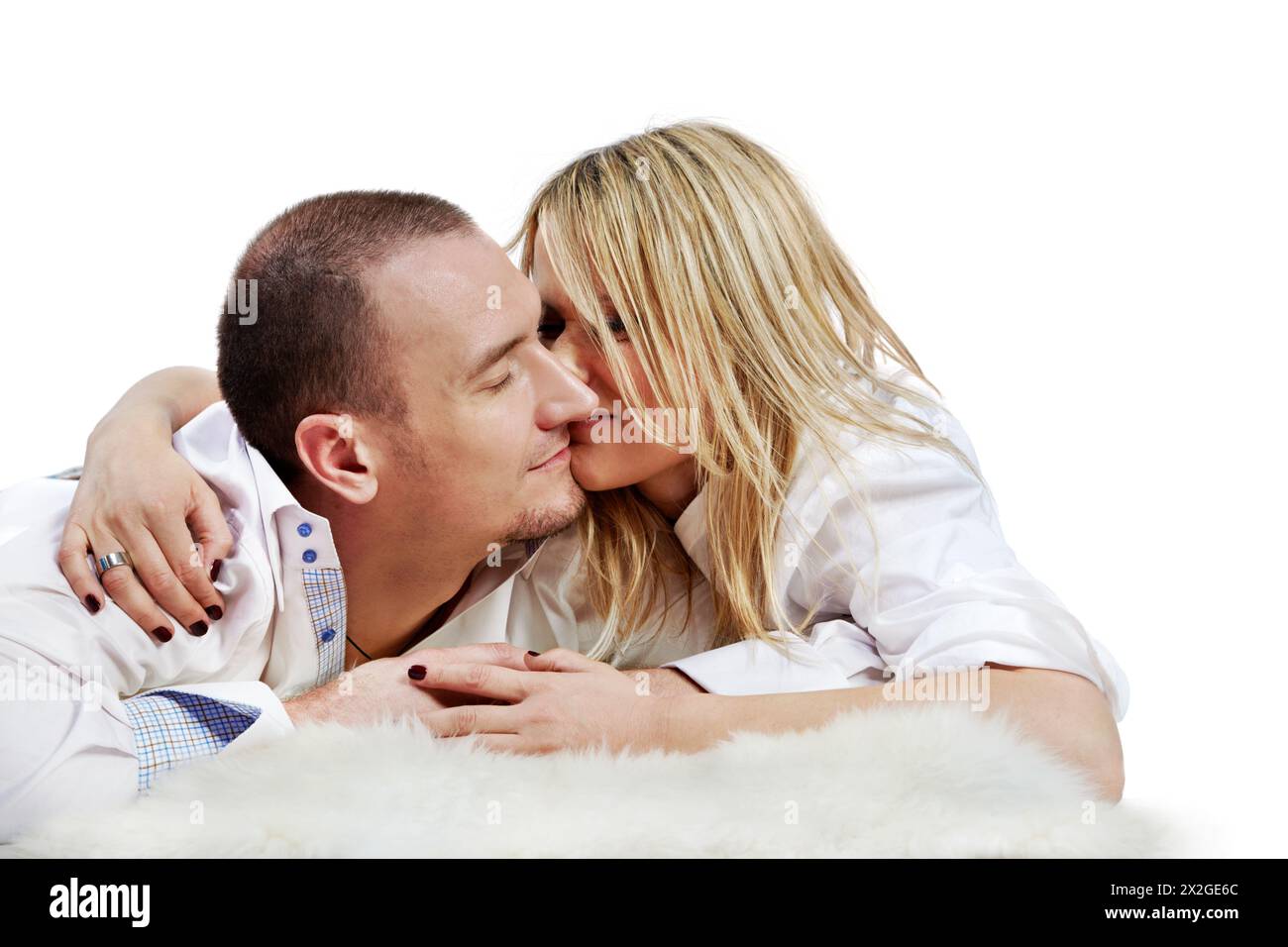 The young pair in white shirts embraces and rubs  their noses, lying on a white fleecy carpet. Stock Photo