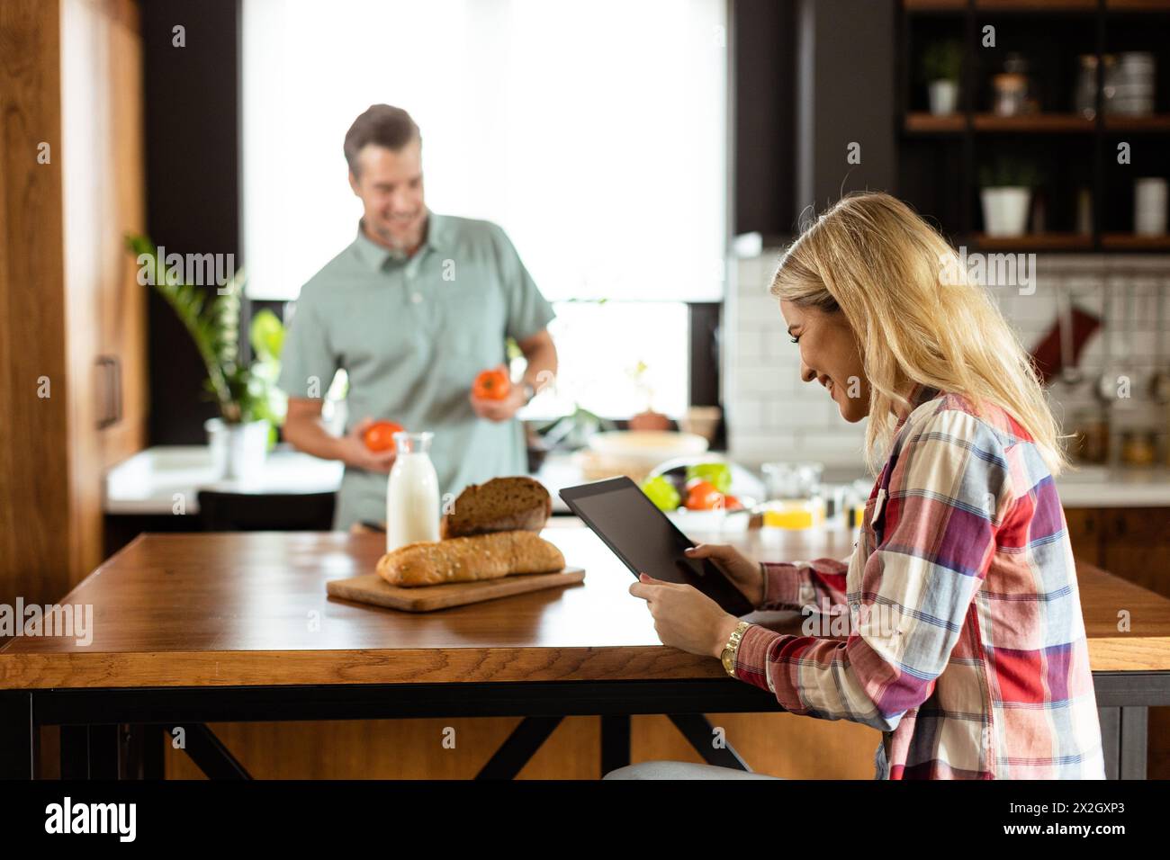 Woman reads from a tablet at the kitchen counter while a man holding an tomatoes smiles at her Stock Photo
